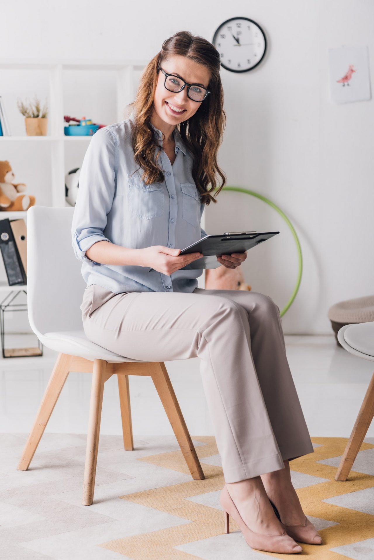 smiling child psychologist with clipboard sitting on chair in office and looking at camera